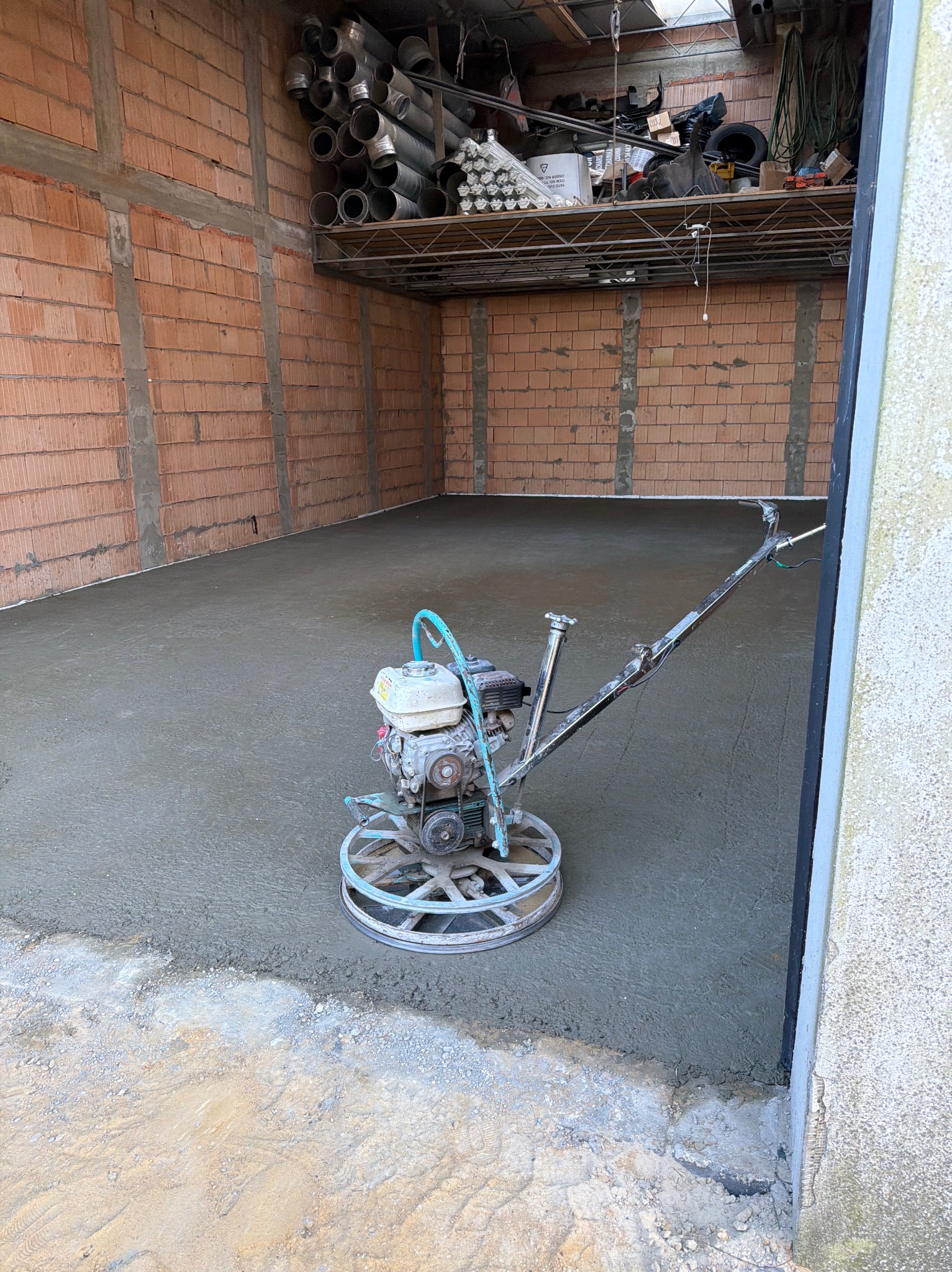 Zacieraczka mechaniczna na świeżo wylanej, szarej posadzce w surowym pomieszczeniu z ceglanymi ścianami i metalową półką z rurami.