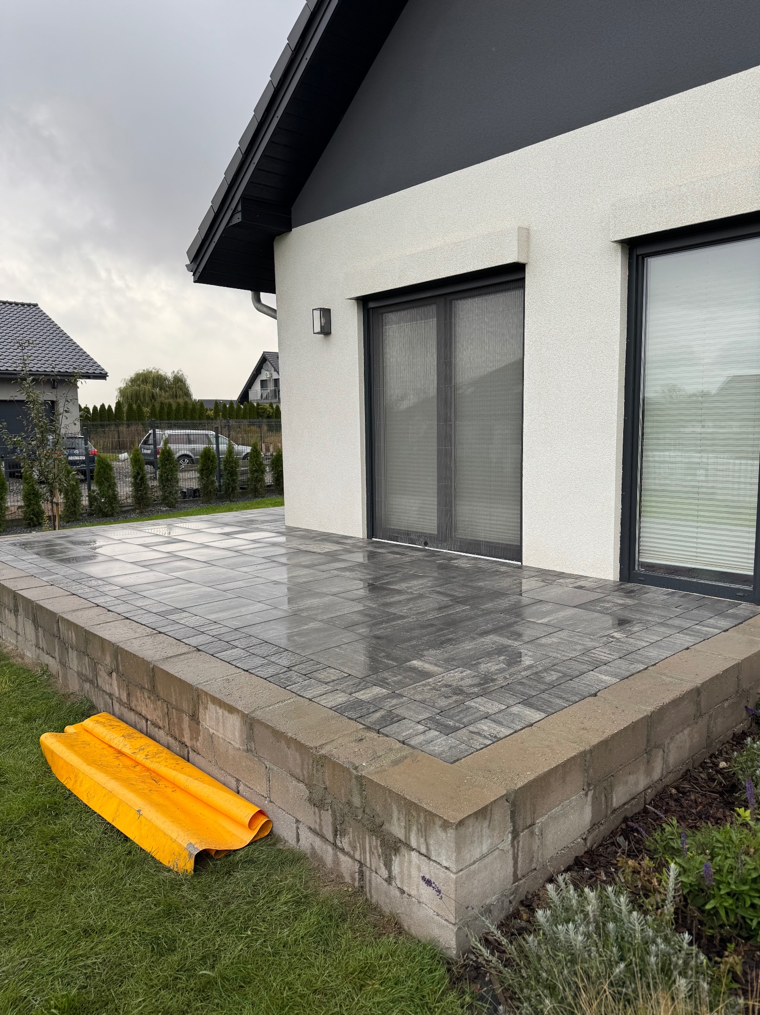 Nowoczesny taras z szarej kostki brukowej przy białym domu z czarnym dachem. Widoczny fragment żółtej osłony na trawie. Pochmurne niebo w tle.