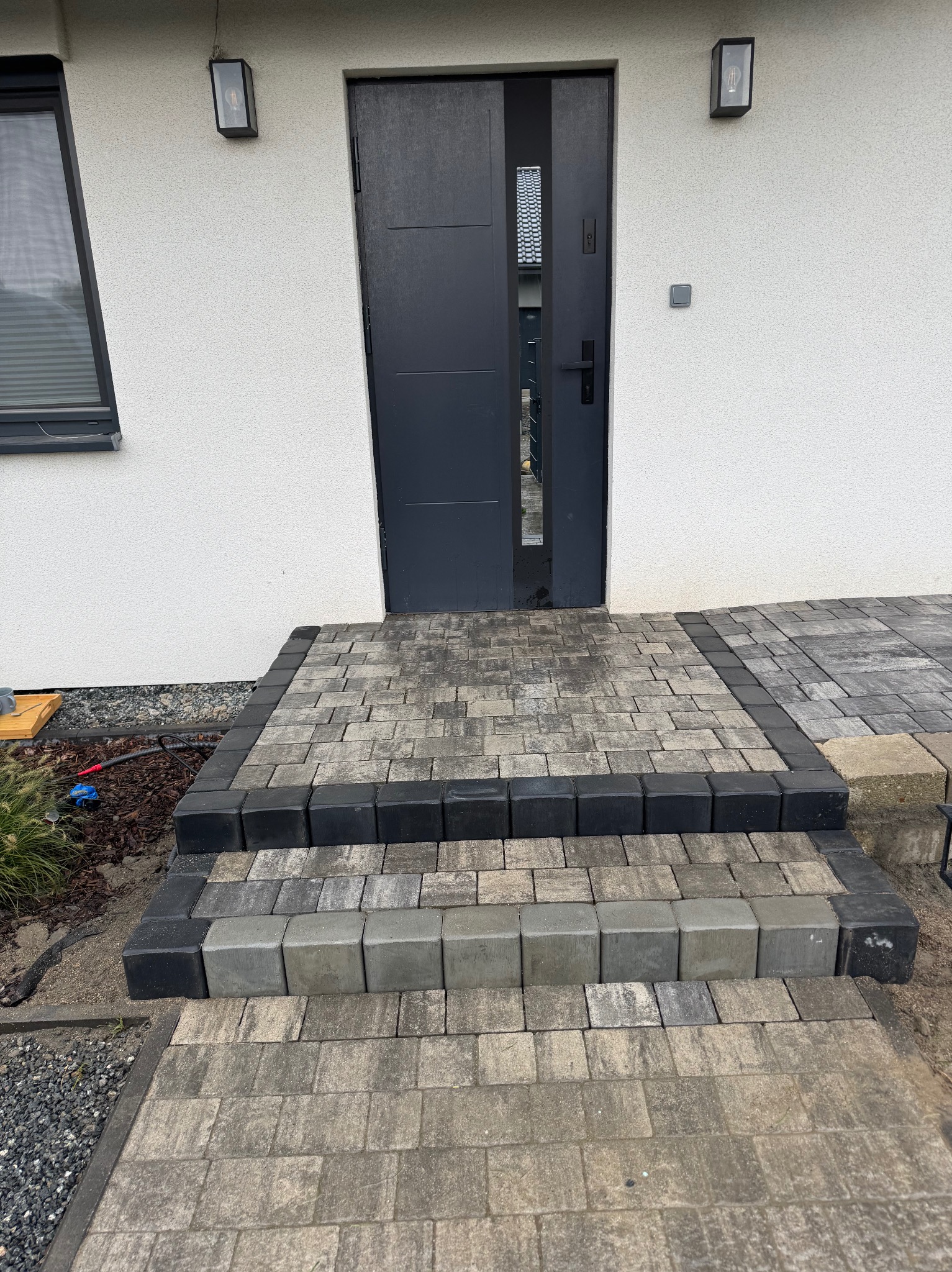 Schody wejściowe wyłożone kostką brukową w odcieniach szarości i czerni, prowadzące do nowoczesnych, grafitowych drzwi wejściowych w białym budynku.