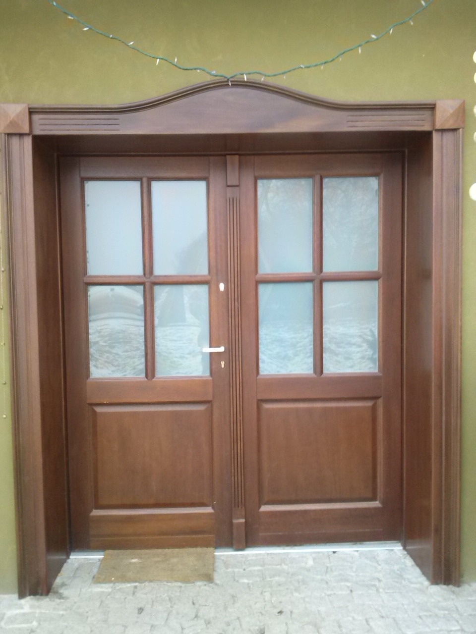 Dwuskrzydłowe, brązowe drzwi drewniane z szybami, ozdobną ramą i małą wycieraczką przed wejściem, oświetlone delikatnym światłem.