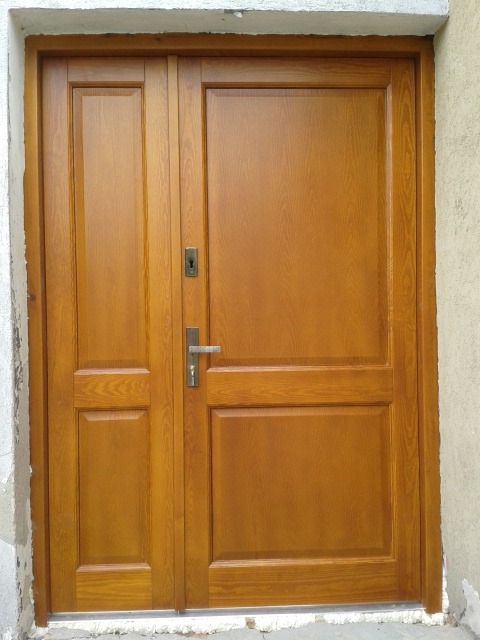 Zewnętrzne, dwuskrzydłowe drzwi wejściowe z drewna o ciepłym, miodowym odcieniu, z widoczną fakturą drewna i metalową klamką, osadzone w surowym, betonowym otworze.