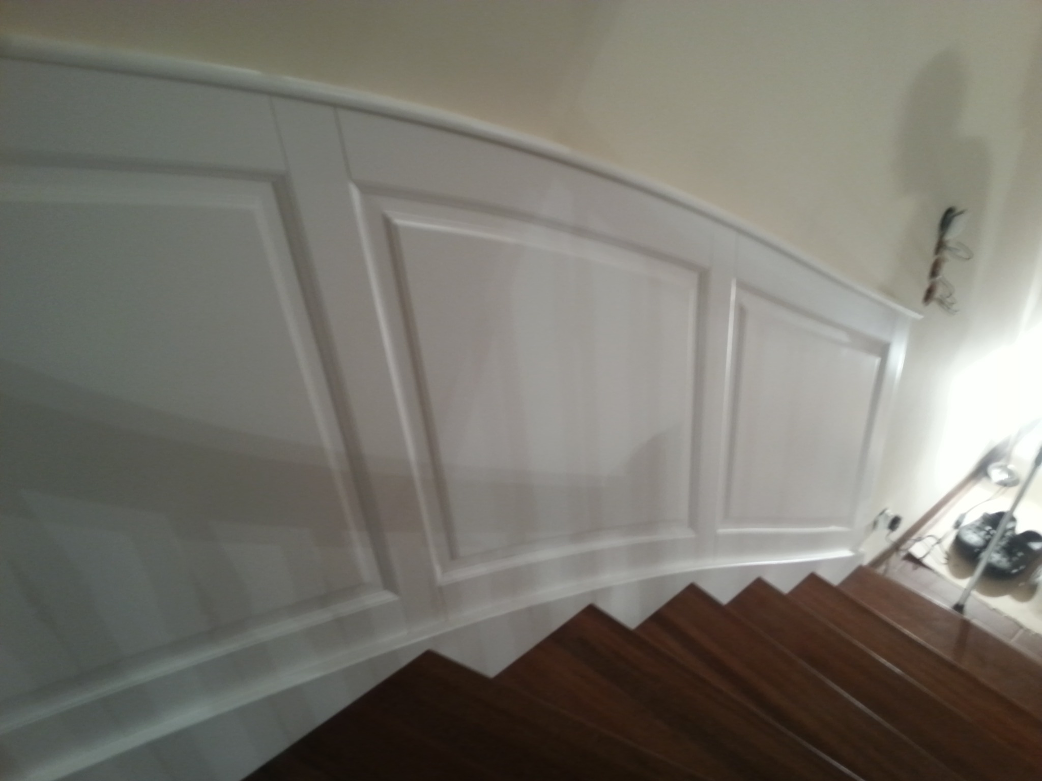 Bieg drewnianych schodów z białą, panelową boazerią ścienną w klasycznym stylu. Ujęcie z góry, podkreślające zakrzywiony kształt schodów i dekoracyjne detale.