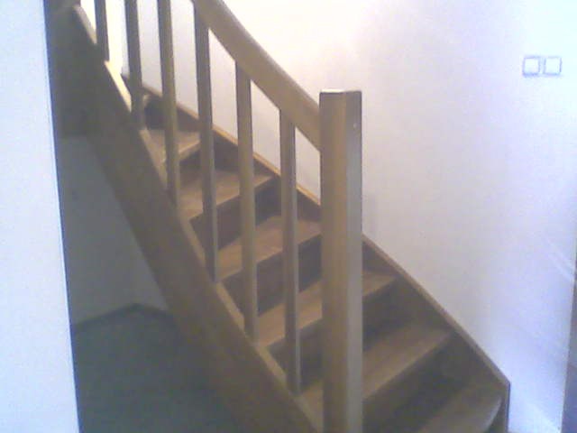 Jasne, drewniane schody z poręczą i pionowymi szczeblami, prowadzące w górę wzdłuż białej ściany z widocznym gniazdkiem elektrycznym. Ujęcie z dołu.