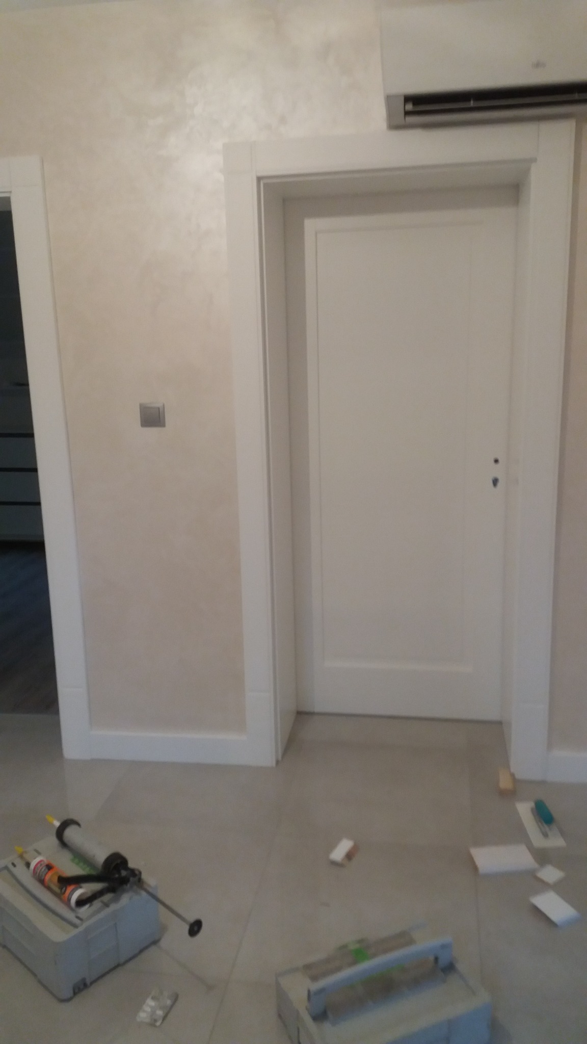 Białe, zamontowane drzwi wewnętrzne z ościeżnicą, widoczne narzędzia montażowe na podłodze. Ściana w odcieniach beżu, klimatyzator nad drzwiami.