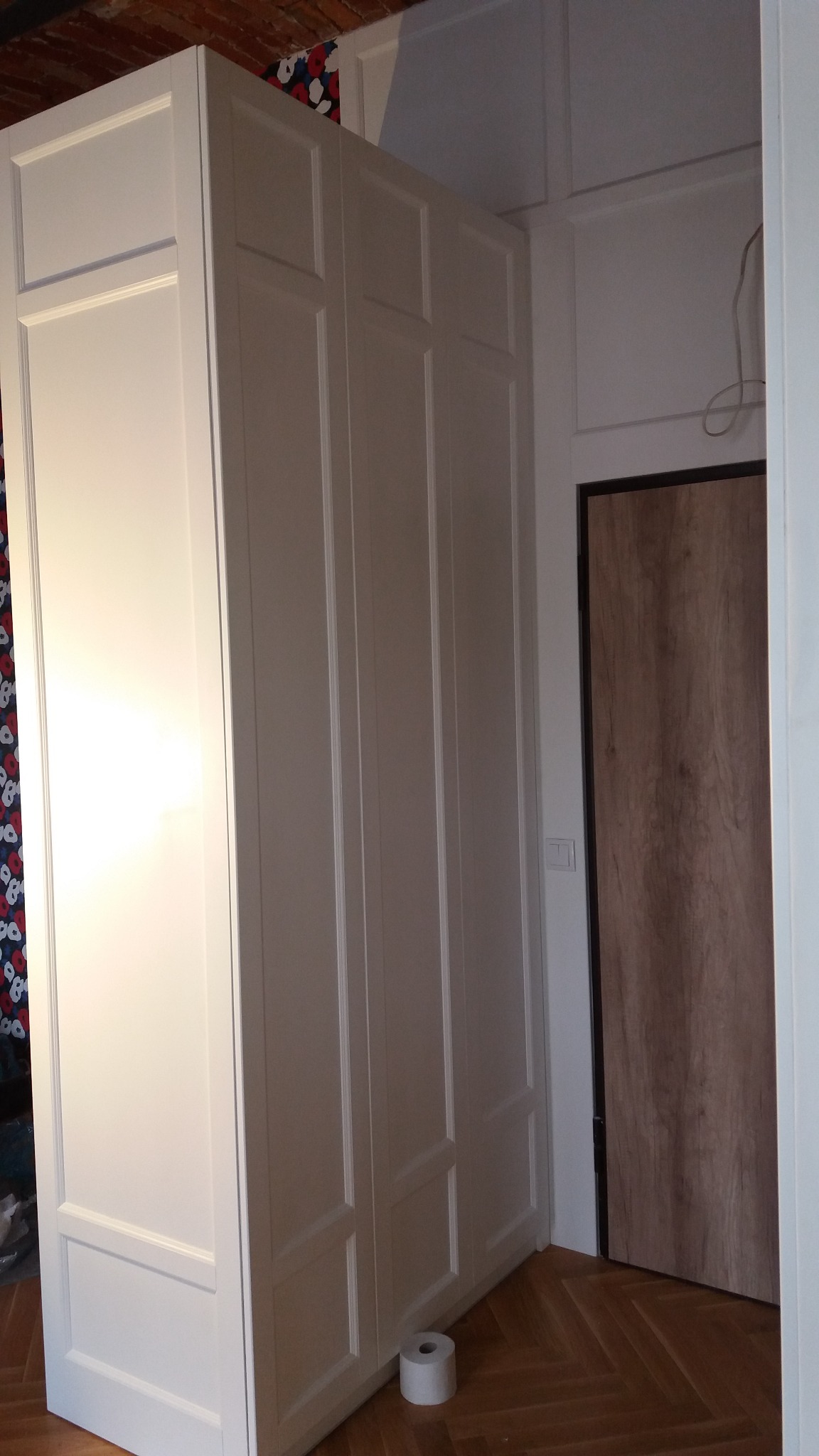 Biała, trzydrzwiowa szafa wnękowa z frezowanymi frontami, ustawiona na parkiecie w jodełkę, z rolką papieru toaletowego u dołu. W tle drzwi z drewnopodobną okleiną.