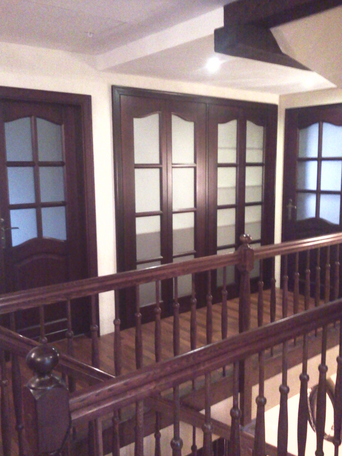 Klatka schodowa z drewnianą balustradą i trzema parami ciemnych, drewnianych drzwi z przeszkleniami. Widok z góry, ciepłe oświetlenie.