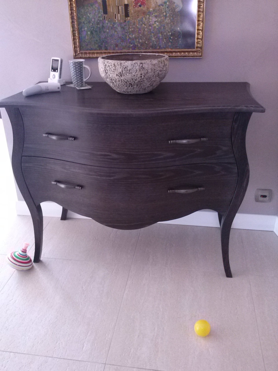 Stylowa, ciemna komoda z dwoma szufladami i giętymi nogami, na blacie dekoracyjna misa, telefon i kubek. W tle obraz w złotej ramie.