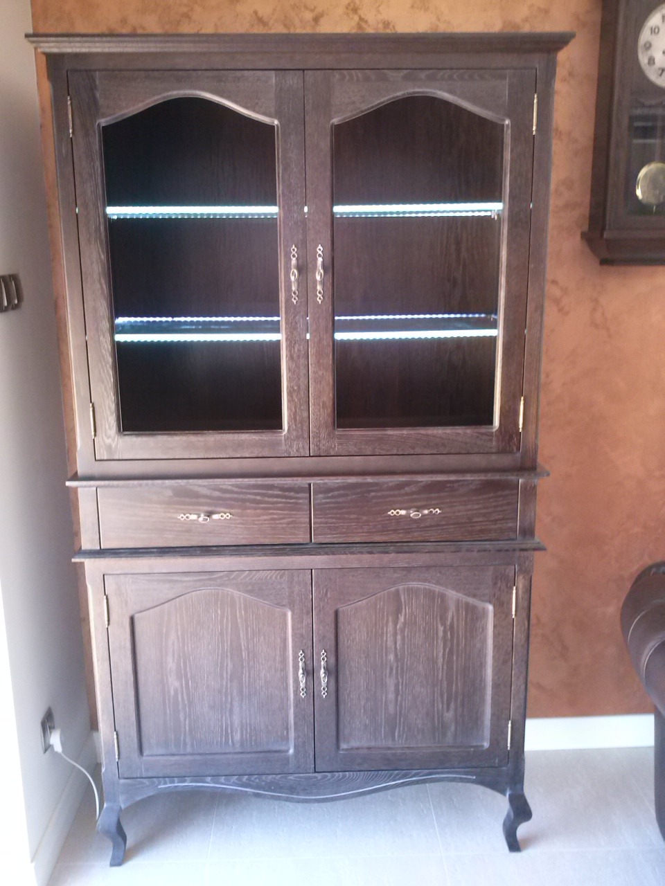 Dwuczęściowa, ciemna witryna z drewna z przeszkloną górną częścią i oświetleniem LED wewnątrz, na tle ściany w ciepłym odcieniu. Klasyczny design z dekoracyjnymi uchwytami.