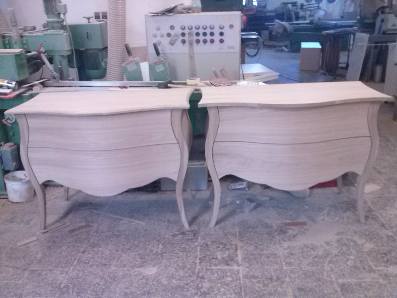 Dwie surowe, drewniane komody w stylu ludwikowskim, bez wykończenia, stojące w warsztacie stolarskim na tle maszyn i narzędzi. Widoczne detale giętych nóg i frontów szuflad.