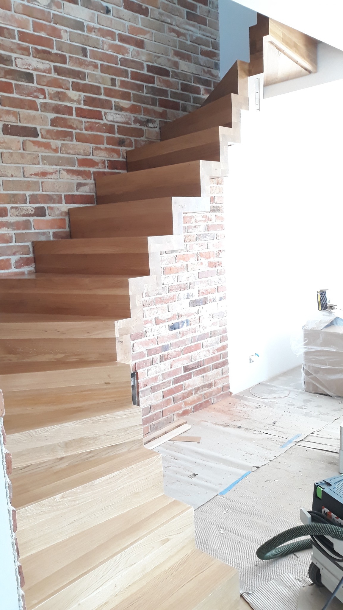 Montaż drewnianych schodów z widoczną ceglaną ścianą. Schody w jasnym odcieniu drewna, surowy stan wykończenia wnętrza, narzędzia pracy widoczne w tle.