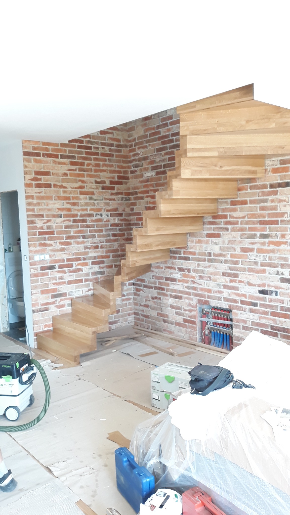 Spiralne schody z jasnego drewna zamontowane przy ceglanej ścianie. Widoczne narzędzia i elementy instalacji grzewczej. Ujęcie z dołu, podkreślające konstrukcję.