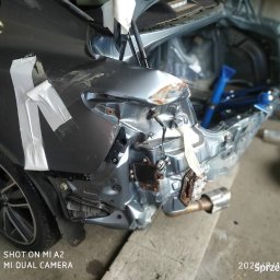 Majcar - Uszkodzony tył szarego samochodu po kolizji, zabezpieczony taśmą. Widoczne wgniecenia, zarysowania i elementy konstrukcyjne. W tle narzędzia warsztatowe.