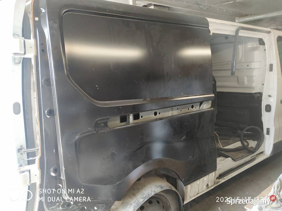 Częściowo zdemontowane, czarne drzwi przesuwne białego busa w warsztacie. Widoczne elementy karoserii i wnętrza pojazdu. Detal z bliska.