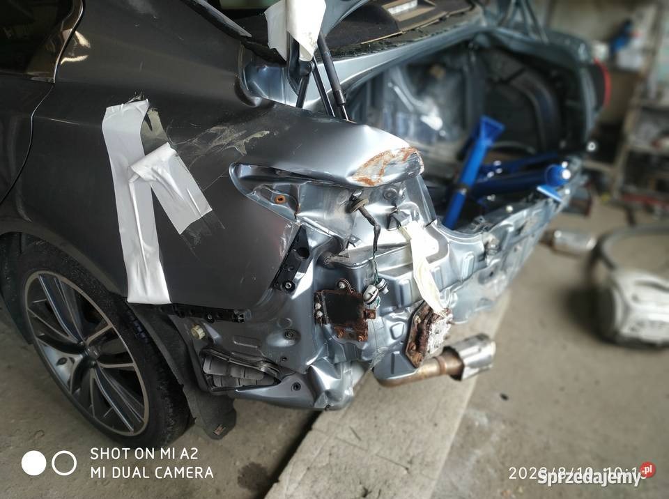 Uszkodzony tył szarego samochodu po kolizji, zabezpieczony taśmą. Widoczne wgniecenia, zarysowania i elementy konstrukcyjne. W tle narzędzia warsztatowe.