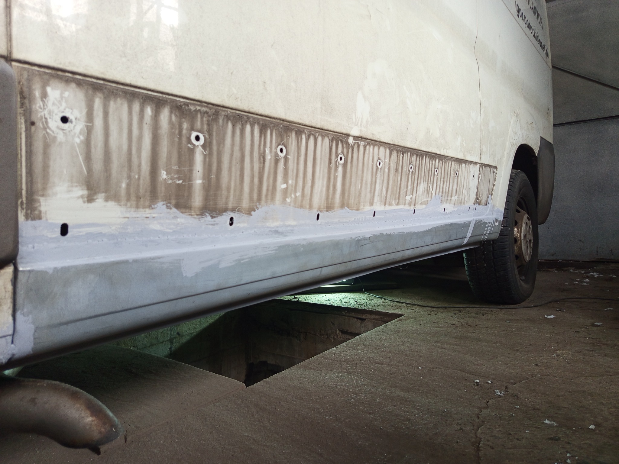 Biały bus na podnośniku w warsztacie, z widocznymi śladami korozji i naprawianym progiem. Widoczne otwory i szpachla na karoserii.