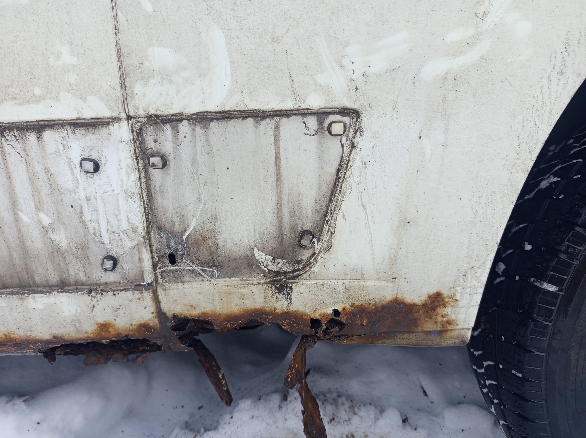 Silna korozja na progu białego samochodu, widoczne dziury i odpadający lakier. Zima, śnieg na dole kadru. Rdza obejmuje znaczną część dolnej krawędzi.
