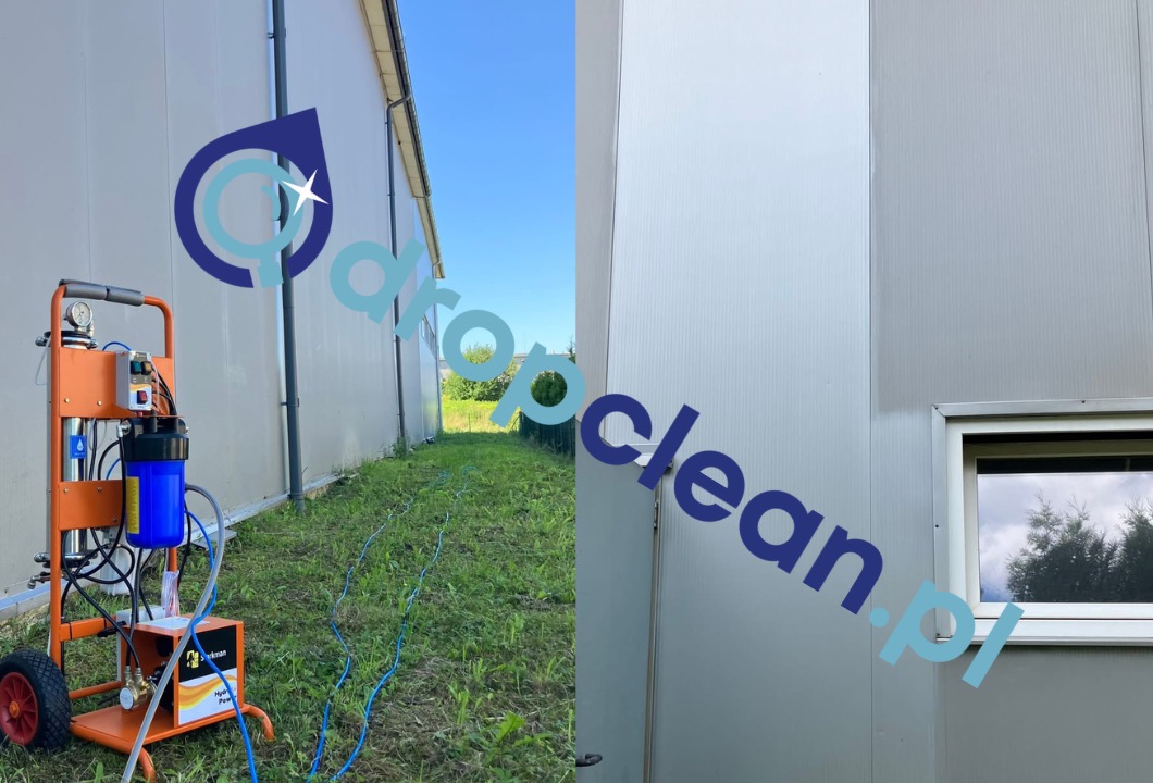 Przenośny agregat ciśnieniowy z niebieskim wężem na trawie, na tle wyczyszczonej, jasnej elewacji hali z oknem. Widoczny fragment elewacji przed i po czyszczeniu.