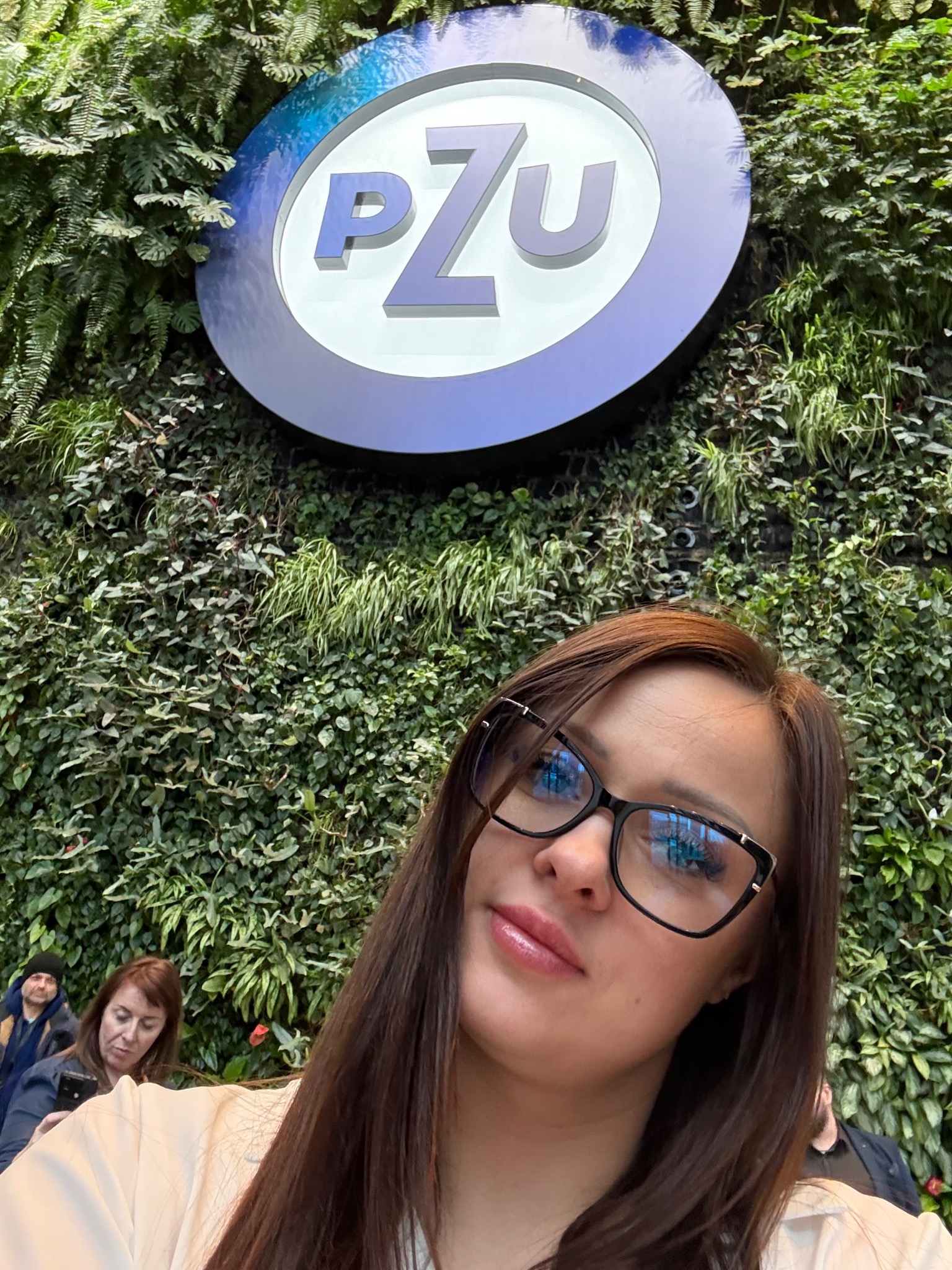 Agentka ubezpieczeniowa na tle ściany z logiem PZU. Kobieta w okularach, uśmiechnięta, z widocznym fragmentem zielonej ściany i logotypu firmy.