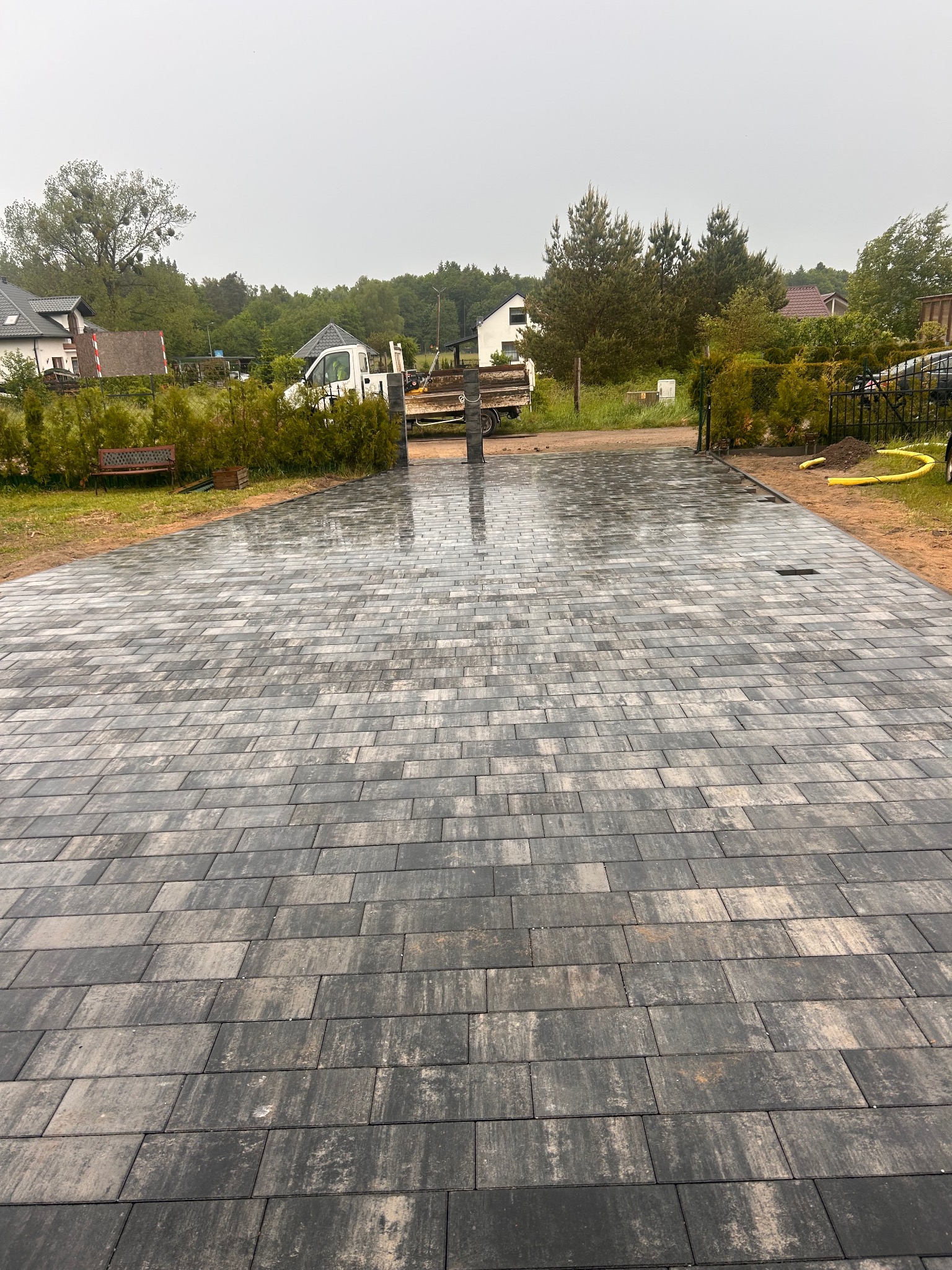 Ułożona, szara kostka brukowa na podjeździe, widok z góry. W tle brama wjazdowa, zieleń i budynki mieszkalne. Lekko mokra nawierzchnia.