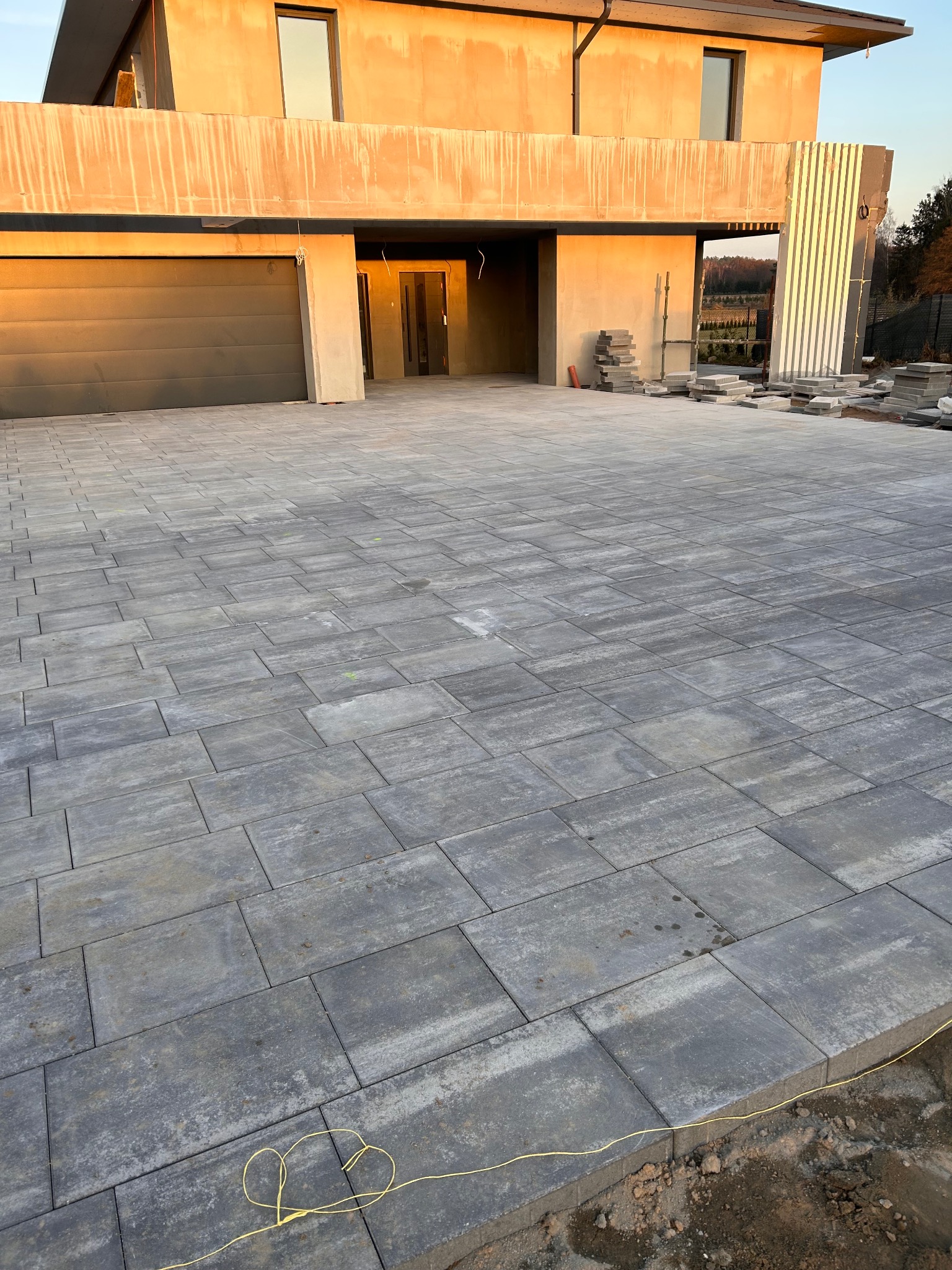 Nowoczesny podjazd z szarej kostki brukowej przed domem w trakcie budowy, widoczne ułożone płyty i przygotowane materiały. Słoneczne popołudnie.