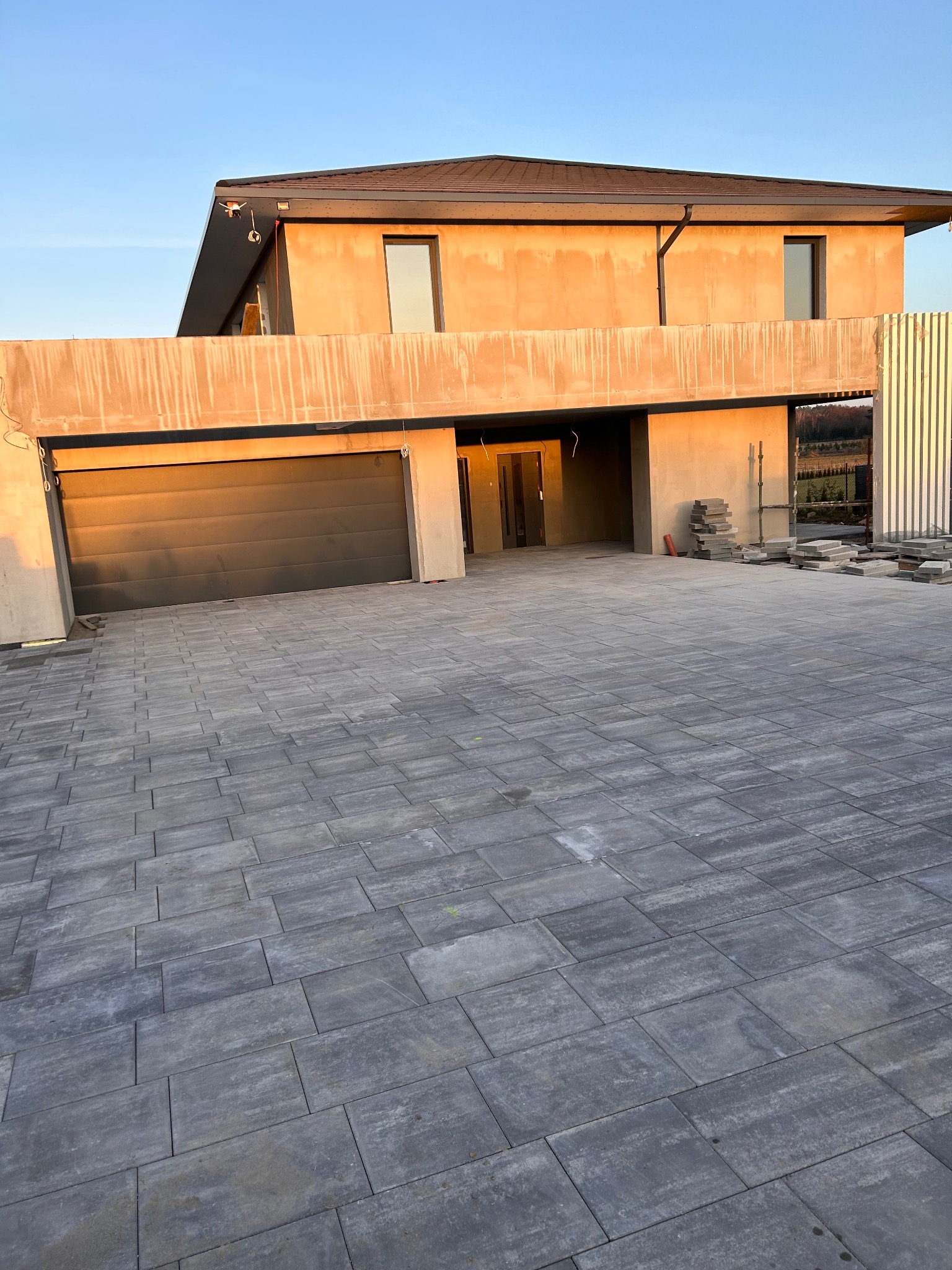 Ułożona szara kostka brukowa na podjeździe przed nowoczesnym domem w surowym stanie. Widoczny garaż z bramą i fragment ogrodzenia.