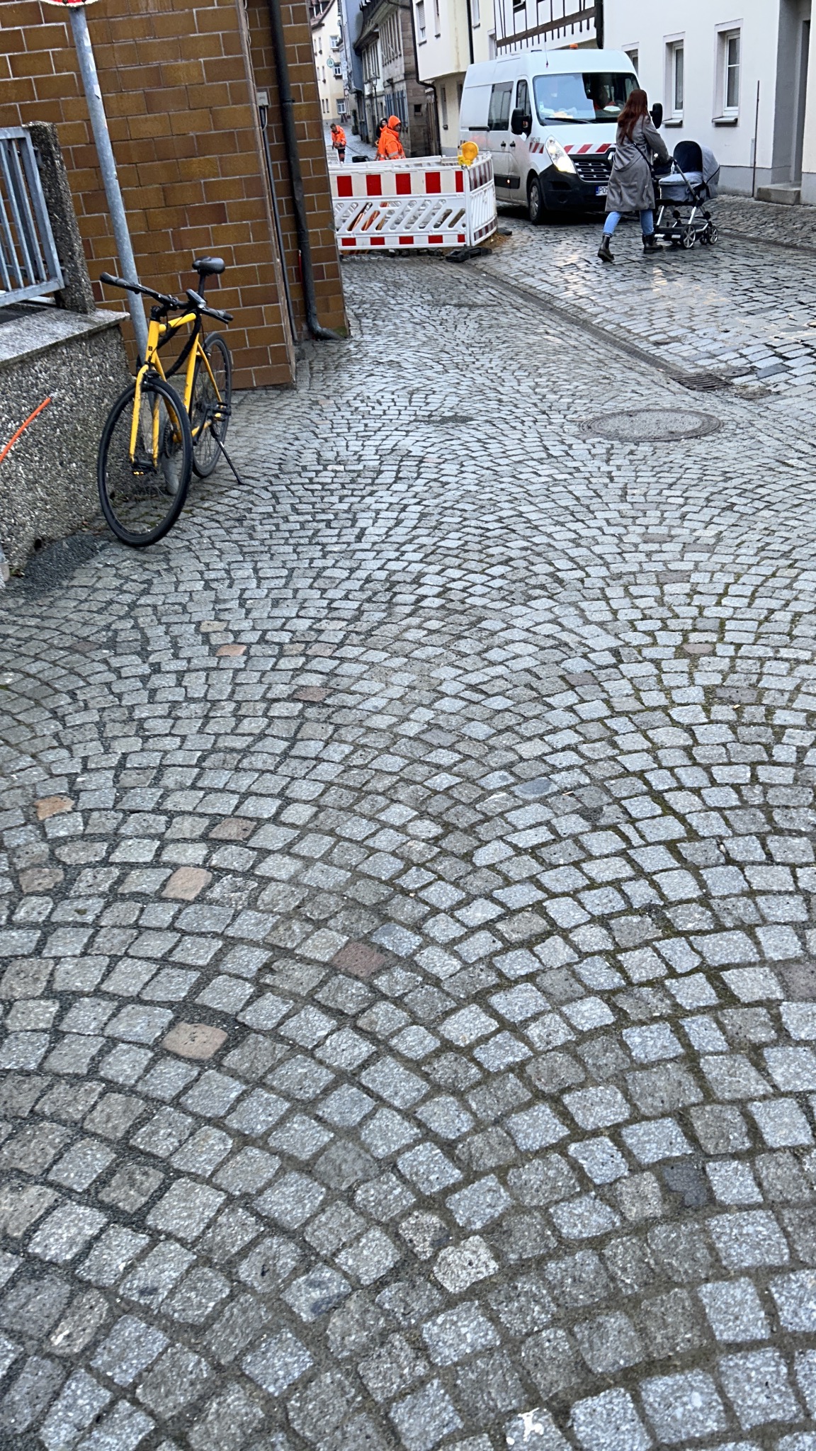 Ulica wyłożona kostką granitową w odcieniach szarości, z żółtym rowerem opartym o budynek. W tle widoczni robotnicy w pomarańczowych strojach i biały samochód dostawczy.