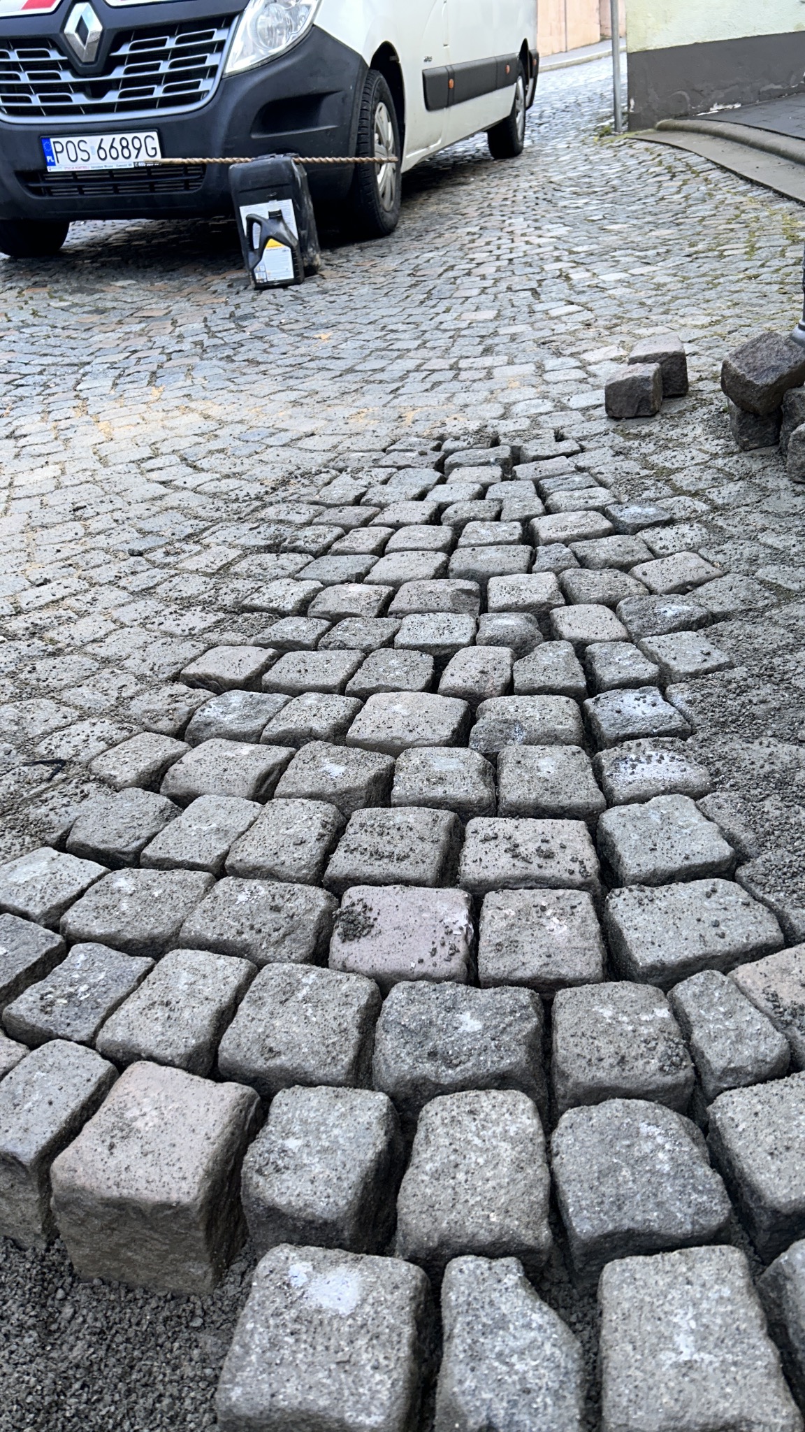 Fragment układanej nawierzchni z kostki granitowej, widoczny samochód dostawczy i kanister z olejem. Częściowo ułożona kostka tworzy wzór w kształcie klina.