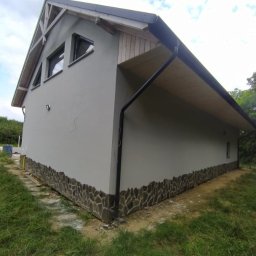 Usługi Budowlane Arkadiusz Jachim - Otynkowany budynek z kamienną podmurówką i drewnianym wykończeniem dachu, widoczny z zewnątrz w słoneczny dzień. Detal: czarna rynna.