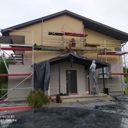 Usługi Budowlane Arkadiusz Jachim - Elewacja domu w trakcie prac wykończeniowych. Widoczne rusztowania, osoby pracujące na wysokości, zabezpieczenia folią i dwa psy siedzące przed wejściem.
