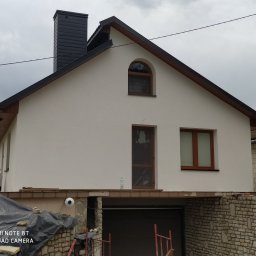 Usługi Budowlane Arkadiusz Jachim - Ocieplony dom jednorodzinny z czarnym kominem i drewnianą stolarką okienną. Widoczne rusztowania i ekipa remontowa. Prace wykończeniowe w toku.