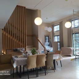 Usługi Budowlane Arkadiusz Jachim - Jasne wnętrze domu w trakcie wykańczania: drewniane schody z ażurową balustradą, marmurowe podłogi, wiszące lampy kule i rusztowanie przy oknie. Widok na jadalnię.