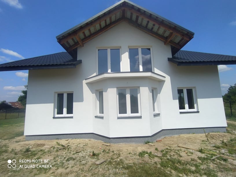 Biały, dwukondygnacyjny dom z ciemnym dachem i jasnymi oknami w słoneczny dzień. Widoczne wykończenie elewacji i otoczenie domu. Ujęcie z dołu.