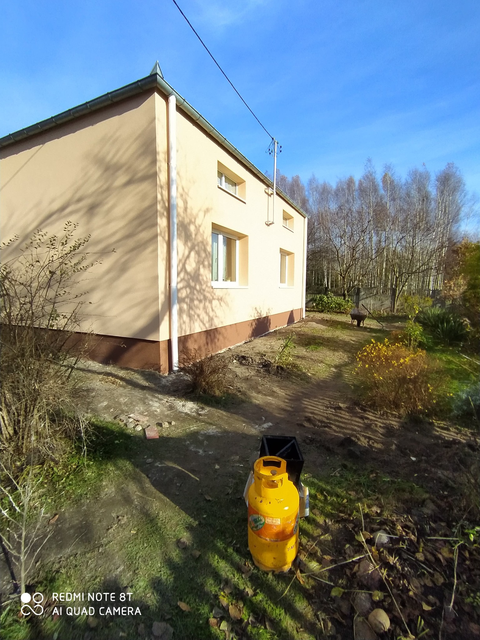 Ocieplony budynek z beżową elewacją i brązowym cokołem, widoczny z zewnątrz w słoneczny dzień. Na pierwszym planie butla gazowa.
