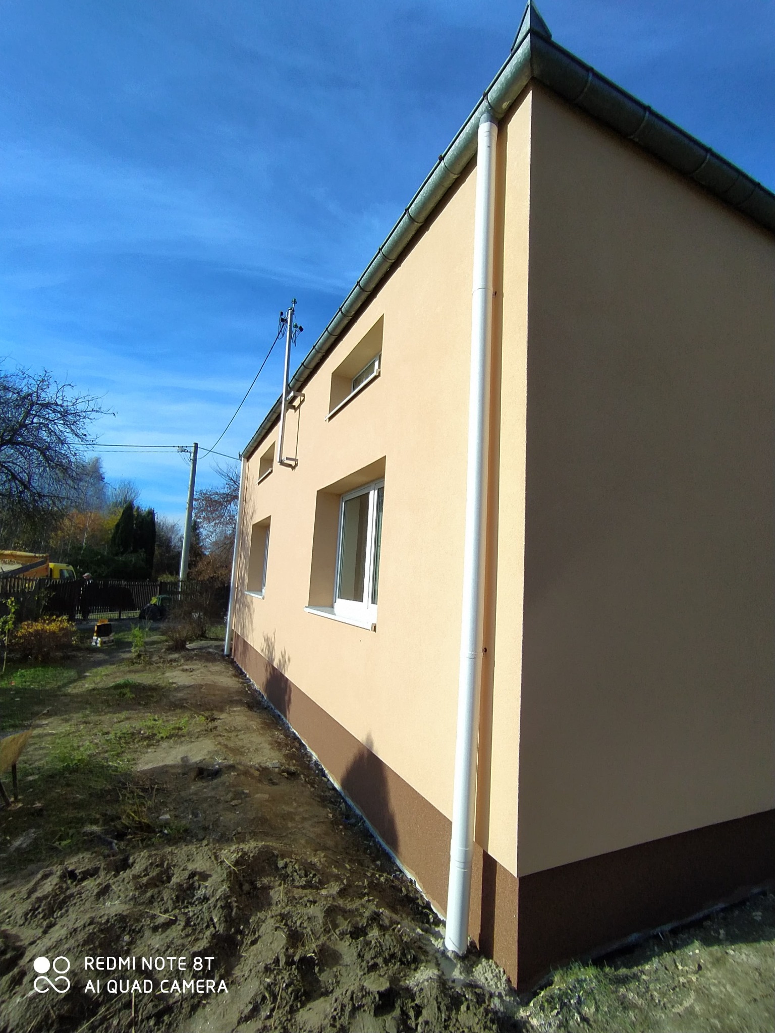 Ocieplony budynek z beżową elewacją i brązowym cokołem, widok z boku na tle błękitnego nieba. Rynna wzdłuż ściany. Widoczne okna i fragment ogrodu.