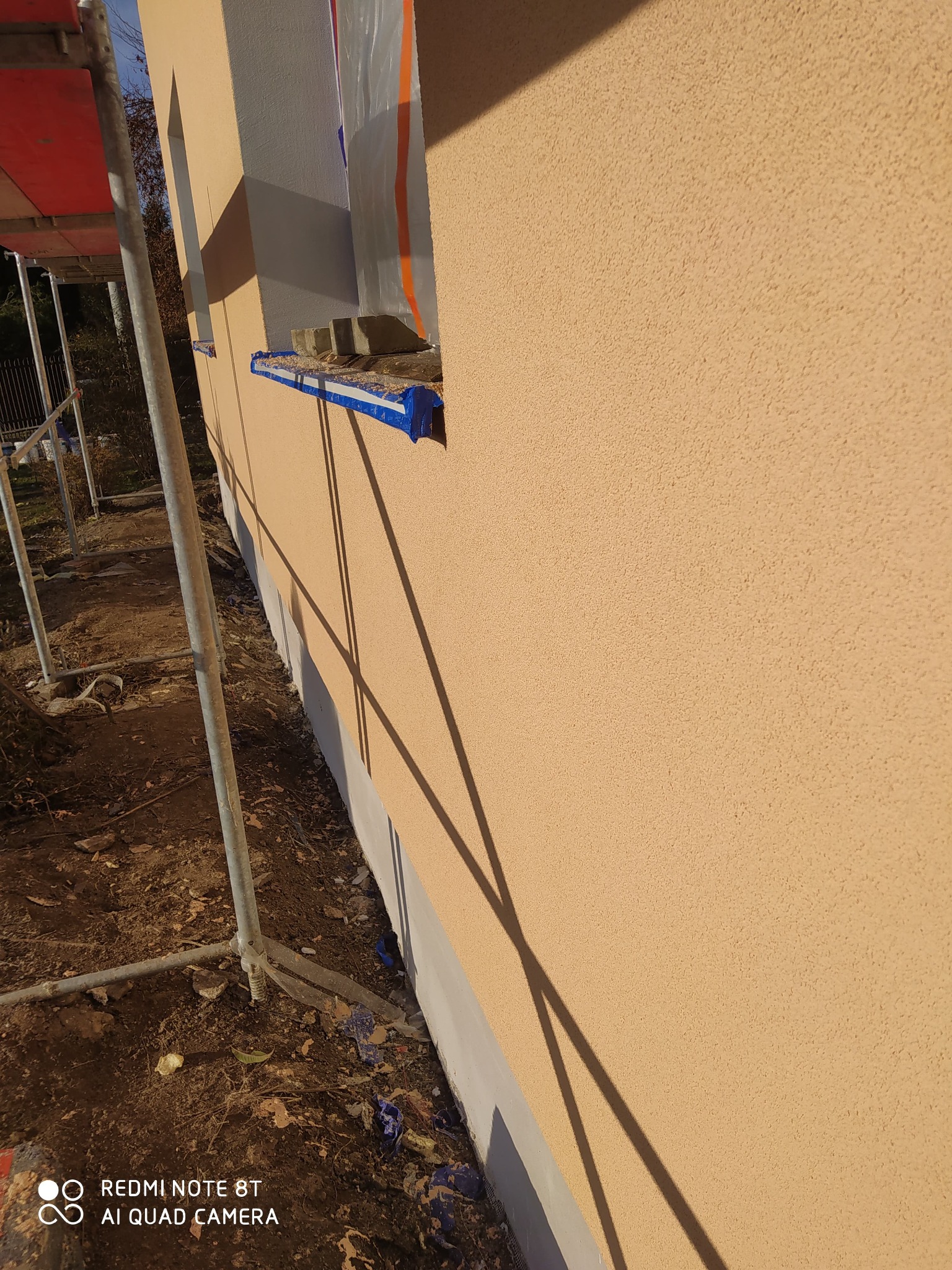 Ściana budynku z nową, beżową elewacją o wyraźnej fakturze, zabezpieczone okno folią i taśmą, widoczne rusztowanie. Detal wykończenia zewnętrznego.