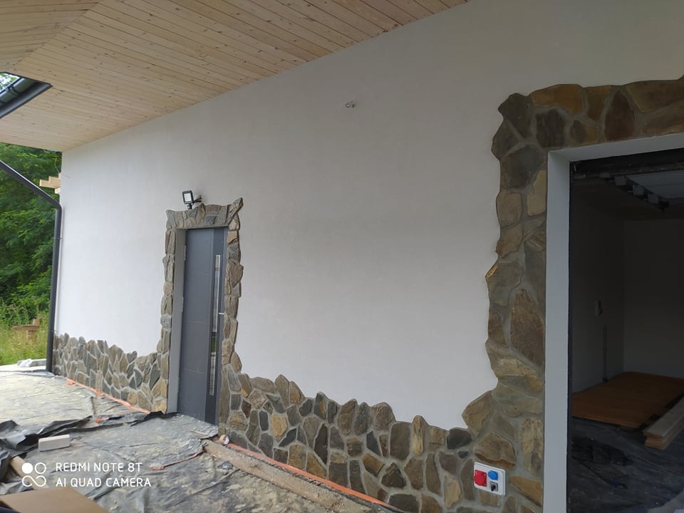 Elewacja domu z dekoracyjnym kamieniem wokół drzwi i u podstawy ściany. Widoczne prace wykończeniowe, folia ochronna na podłożu. Częściowo widoczny drewniany sufit.