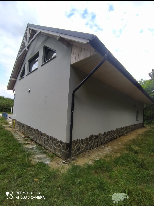 Otynkowany budynek z kamienną podmurówką i drewnianym wykończeniem dachu, widoczny z zewnątrz w słoneczny dzień. Detal: czarna rynna.