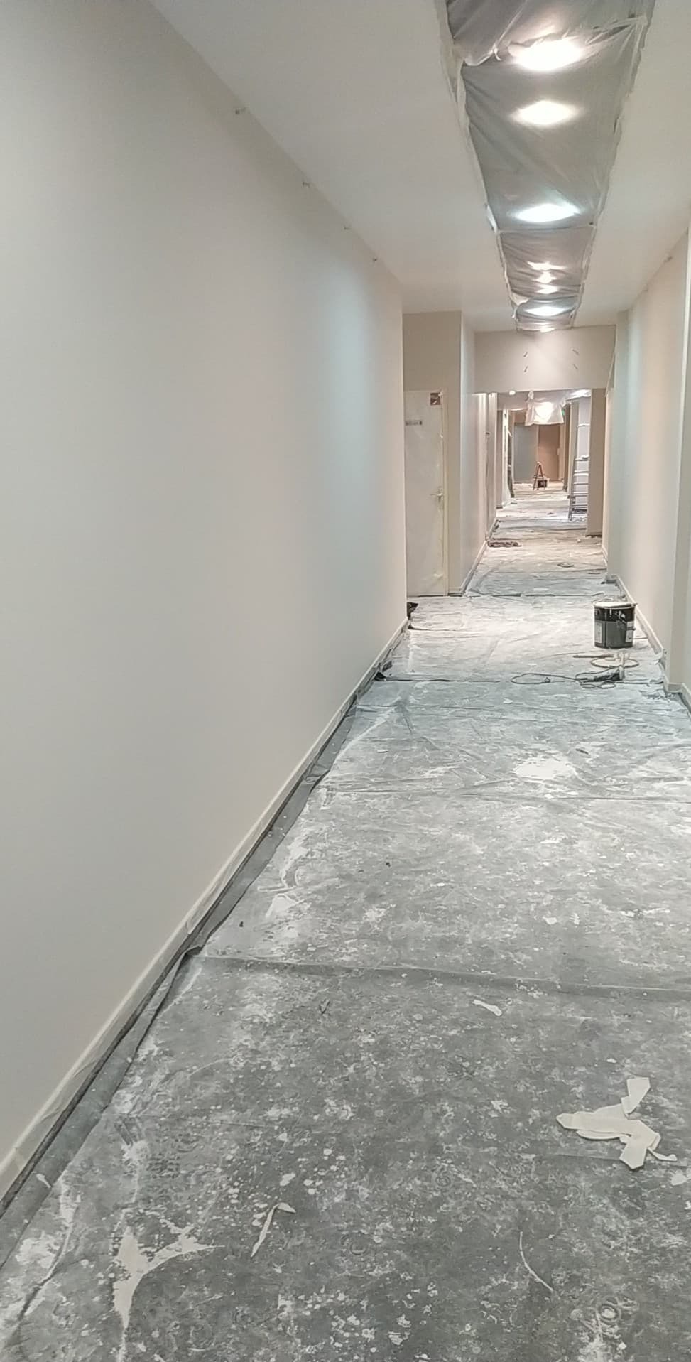 Korytarz w trakcie malowania: świeżo pomalowane ściany w jasnym odcieniu, podłoga zabezpieczona folią malarską, widoczny kubeł z farbą i oświetlenie sufitowe.