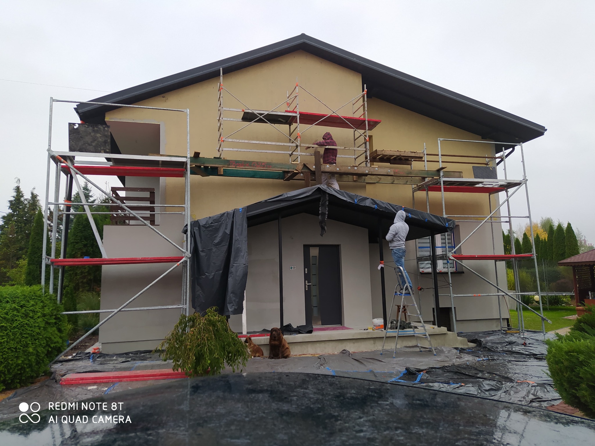 Elewacja domu w trakcie prac wykończeniowych. Widoczne rusztowania, osoby pracujące na wysokości, zabezpieczenia folią i dwa psy siedzące przed wejściem.