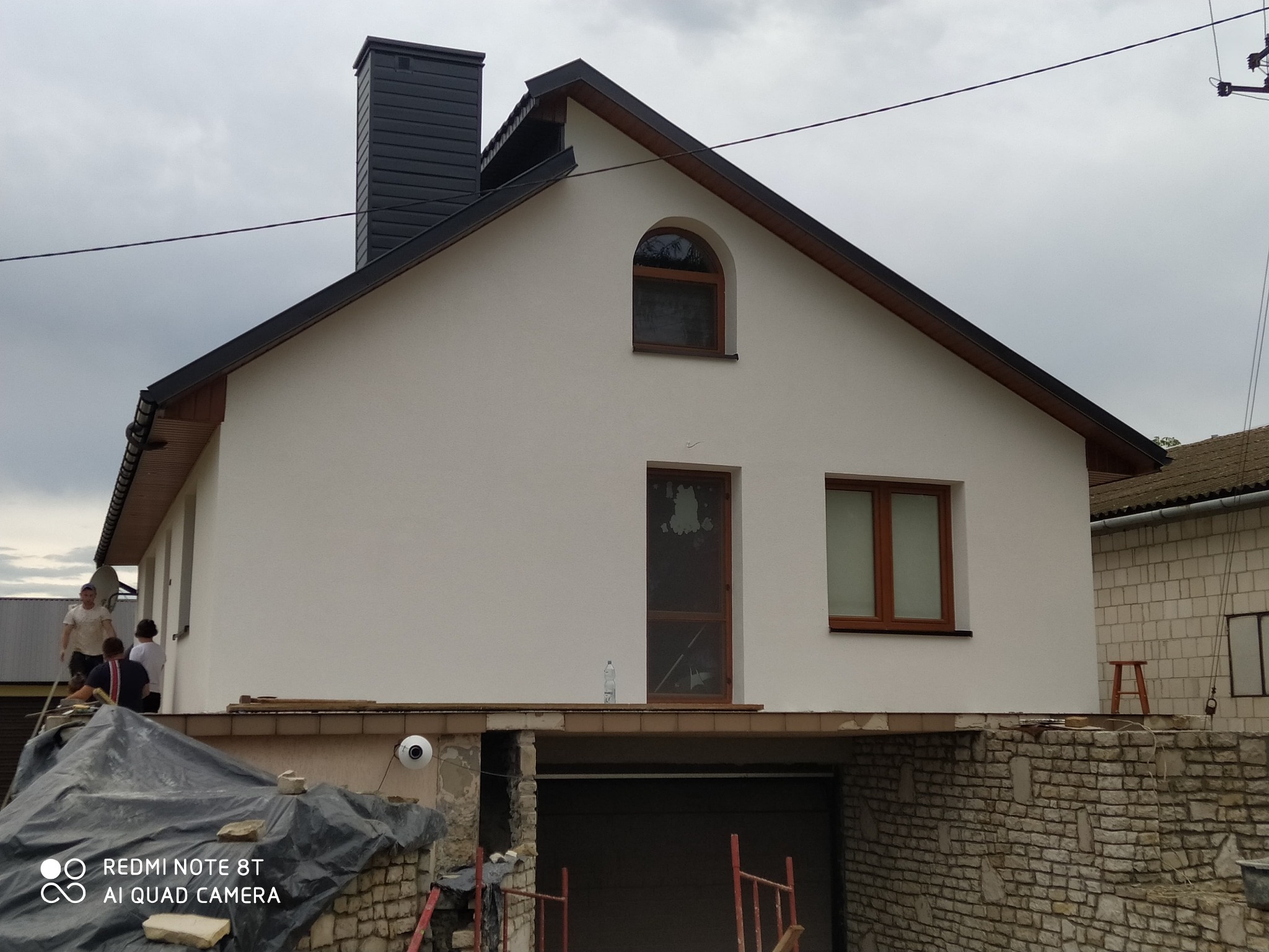 Ocieplony dom jednorodzinny z czarnym kominem i drewnianą stolarką okienną. Widoczne rusztowania i ekipa remontowa. Prace wykończeniowe w toku.