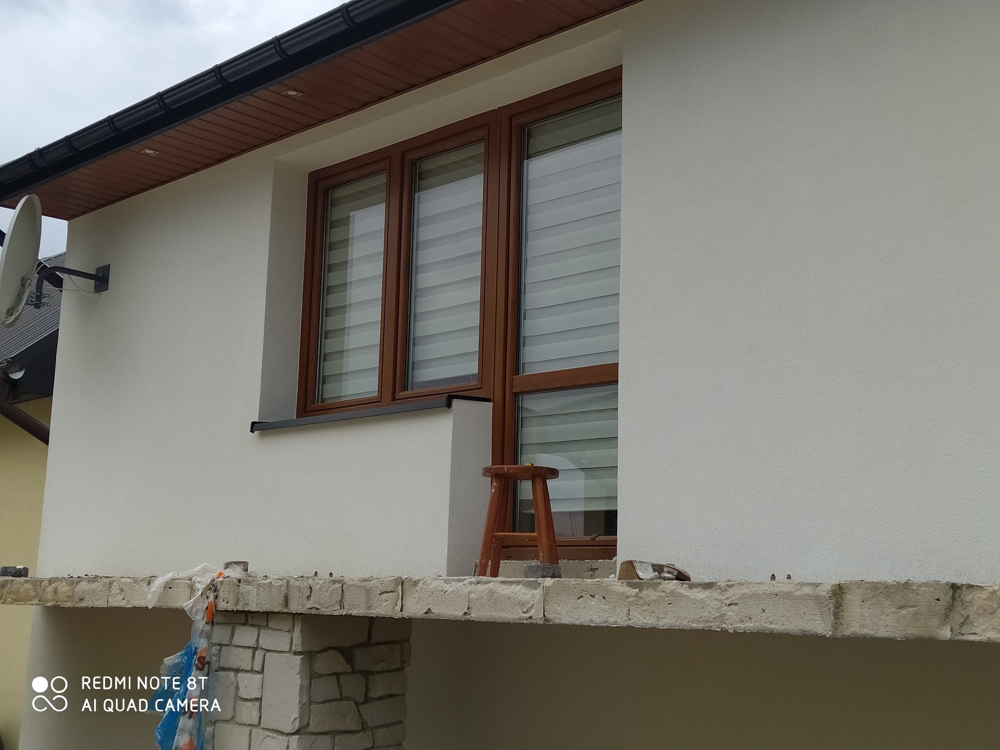Biała elewacja domu z brązowymi oknami i roletami. Widoczny fragment balkonu z ceglaną podstawą i drewniany stołek. Antena satelitarna na ścianie.