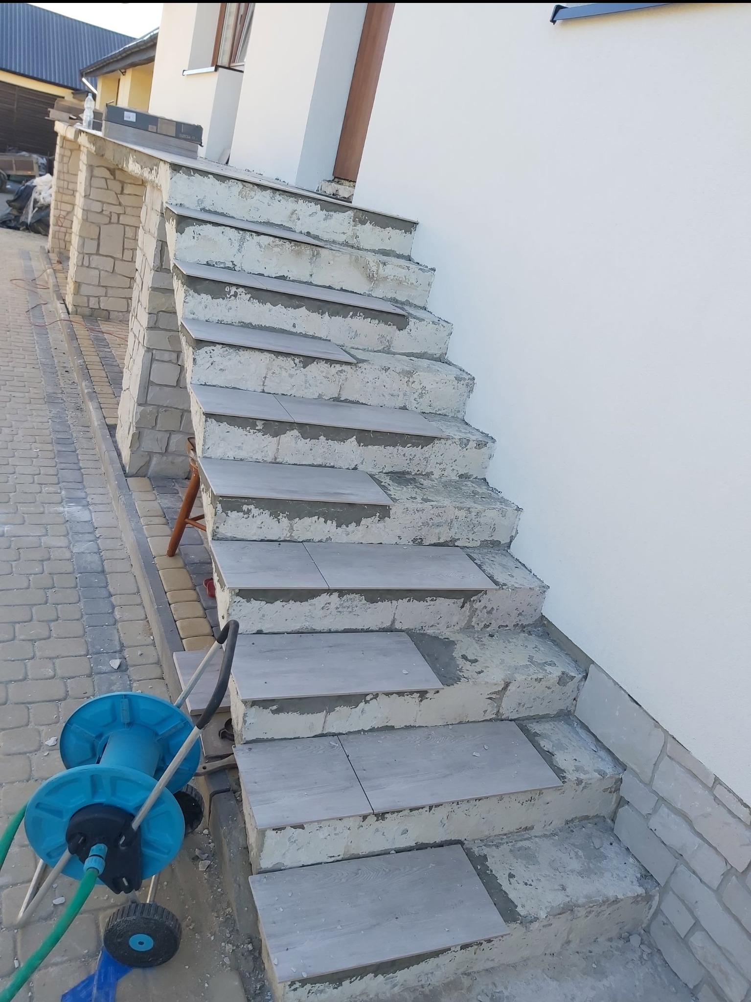 Układanie płytek na schodach zewnętrznych domu. Widoczne niedokończone prace, szare płytki i niebieski zwijak do węża ogrodowego. Ujęcie z dołu, z boku.