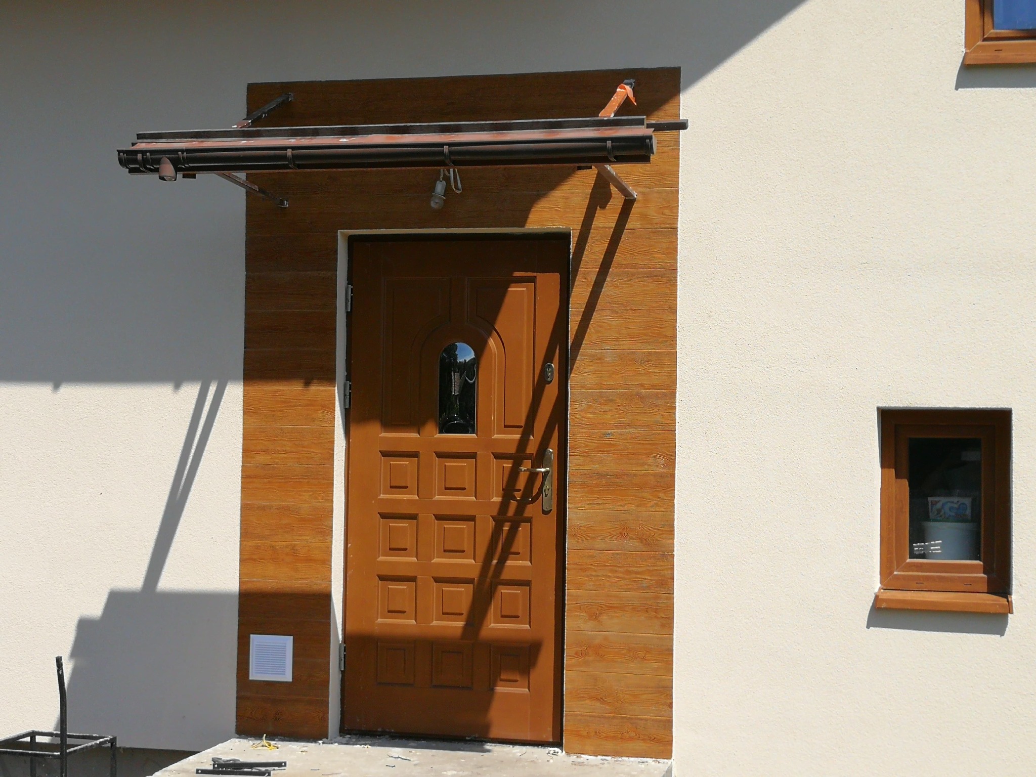 Brązowe drzwi wejściowe z drewnopodobną obudową i zadaszeniem, oświetlone słońcem, z małym oknem po prawej stronie. Widoczne narzędzia i elementy montażowe.