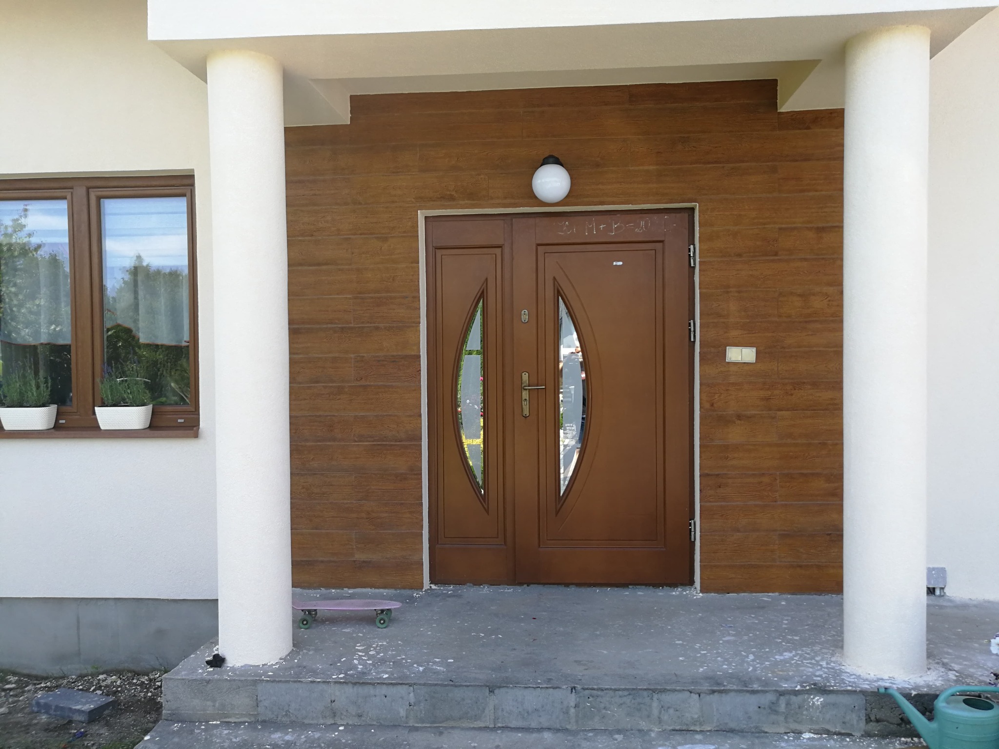 Wejście do domu z brązowymi drzwiami i drewnopodobną elewacją, flankowane białymi kolumnami. Widoczny fragment okna z kwiatami i dziecięca deskorolka.