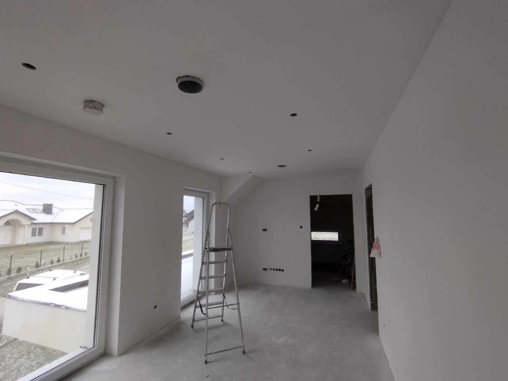 Białe, świeżo pomalowane wnętrze z drabiną, widok na okno i otwór w suficie. Minimalistyczny design, surowy stan wykończenia. Widok na zewnątrz z ośnieżonym krajobrazem.