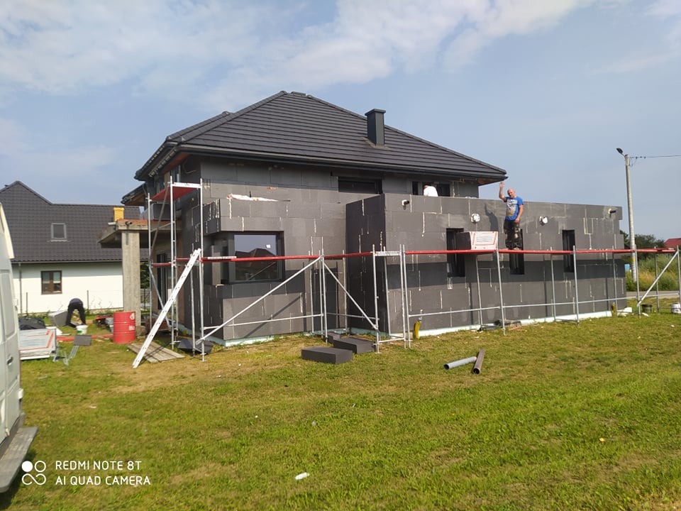 Dom jednorodzinny w trakcie ocieplania styropianem, widoczny pracownik na rusztowaniu, fragment trawnika i nieba. Budynek z ciemną dachówką.