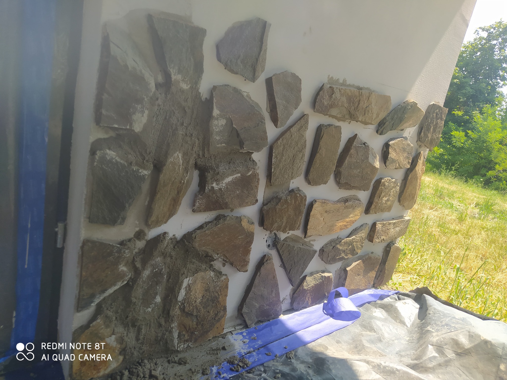 Fragment ściany z kamienia naturalnego w trakcie montażu, widoczne zaprawa i zabezpieczenie podłoża folią. Praca w toku, zieleń w tle.