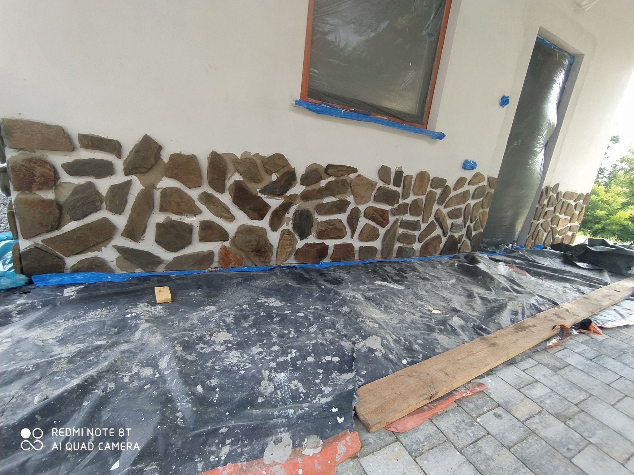 Układanie kamienia elewacyjnego na ścianie budynku. Częściowo wykonana praca, widoczne zabezpieczenia okna i drzwi folią. Na dole rozłożona folia ochronna.