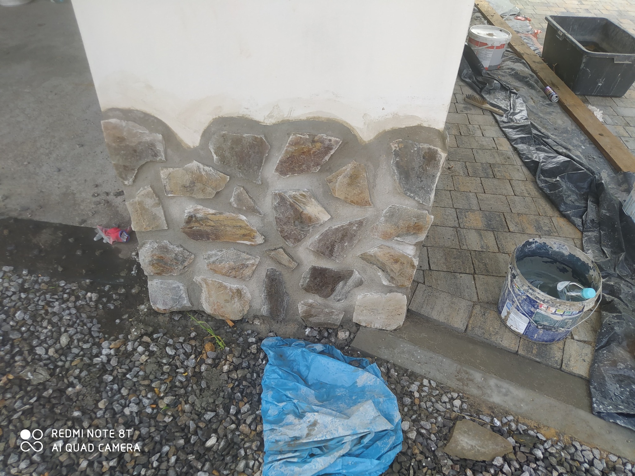 Fragment ściany z dekoracyjnym wykończeniem z kamienia naturalnego, w trakcie montażu. Widoczne narzędzia i materiały budowlane. Częściowo ukończona elewacja.