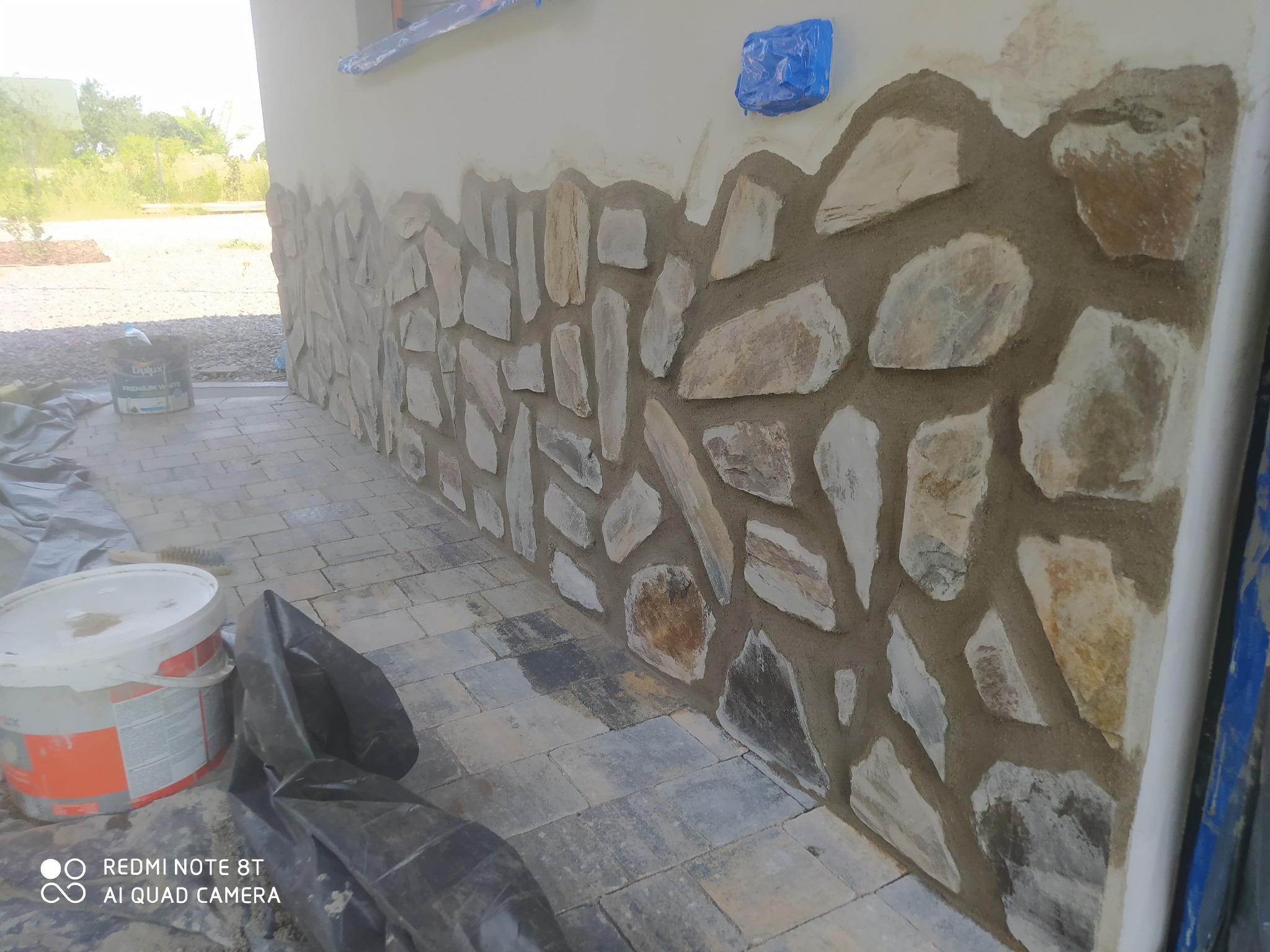 Ściana budynku w trakcie wykończenia kamieniem naturalnym, widoczne narzędzia i materiały budowlane. Prace wykończeniowe na zewnątrz budynku.